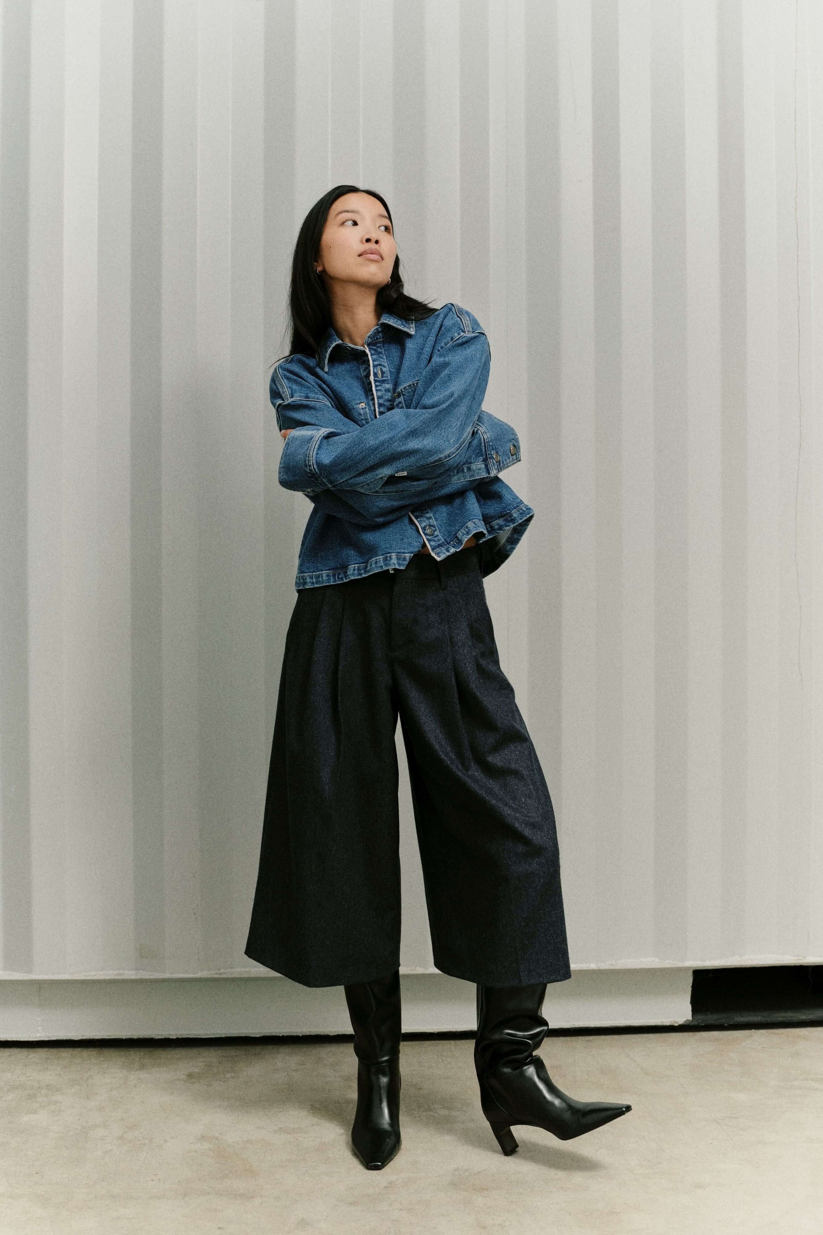Frau in blauer Jeansjacke, dunkler Culotte mit weitem Bein und schwarzen Stiefeln vor gestreiftem Hintergrund.