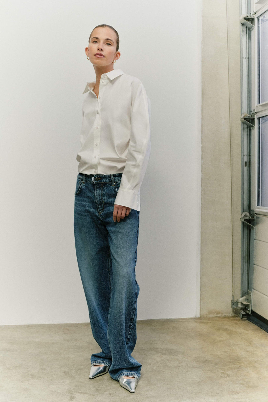 Eine Frau in einer klassischen weißen Bluse, einer blauen Jeans und silbernen, spitzen Schuhen.