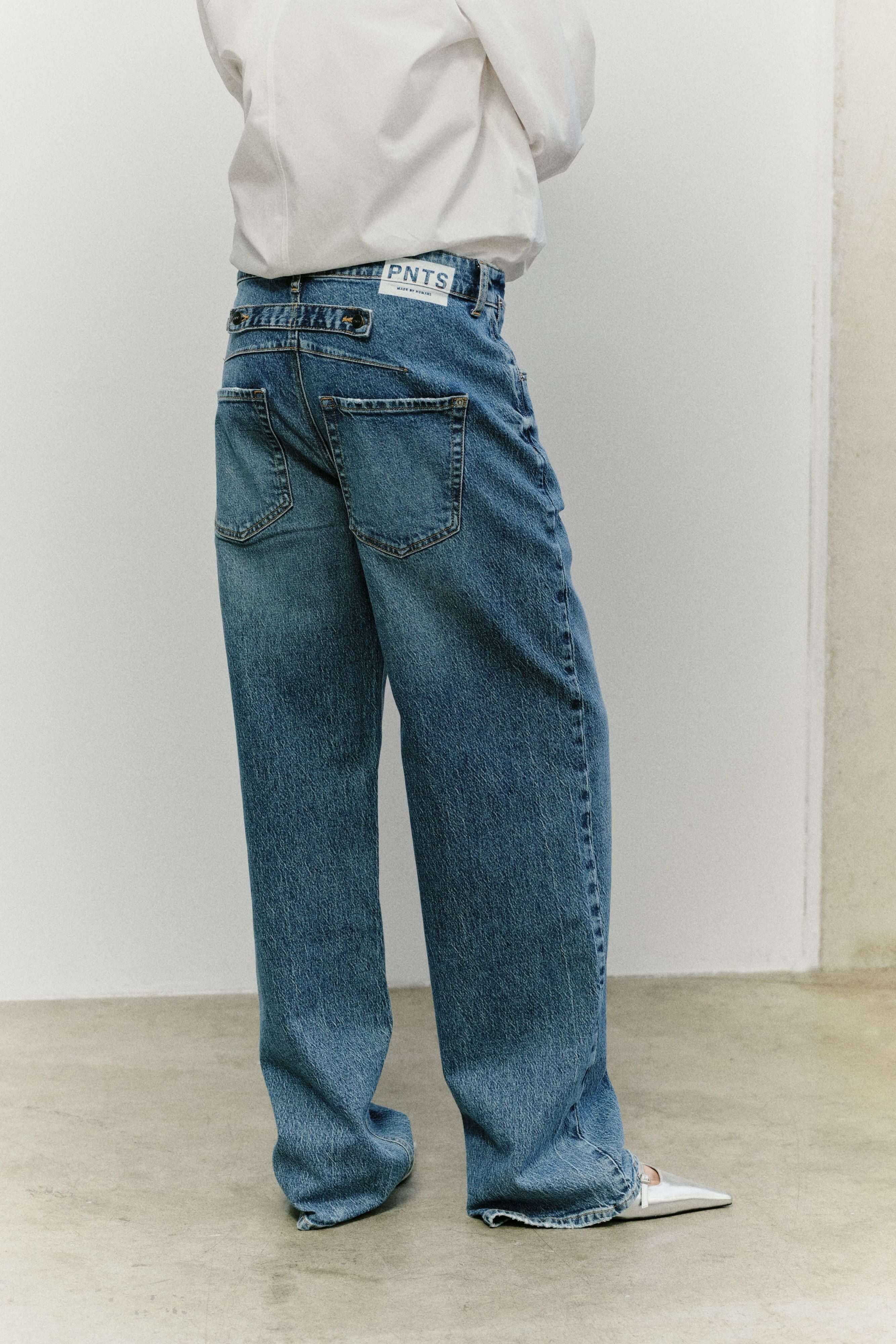 Rückansicht einer blauen Jeans mit PNTS-Label am Bund und silbernen, spitzen Schuhen.