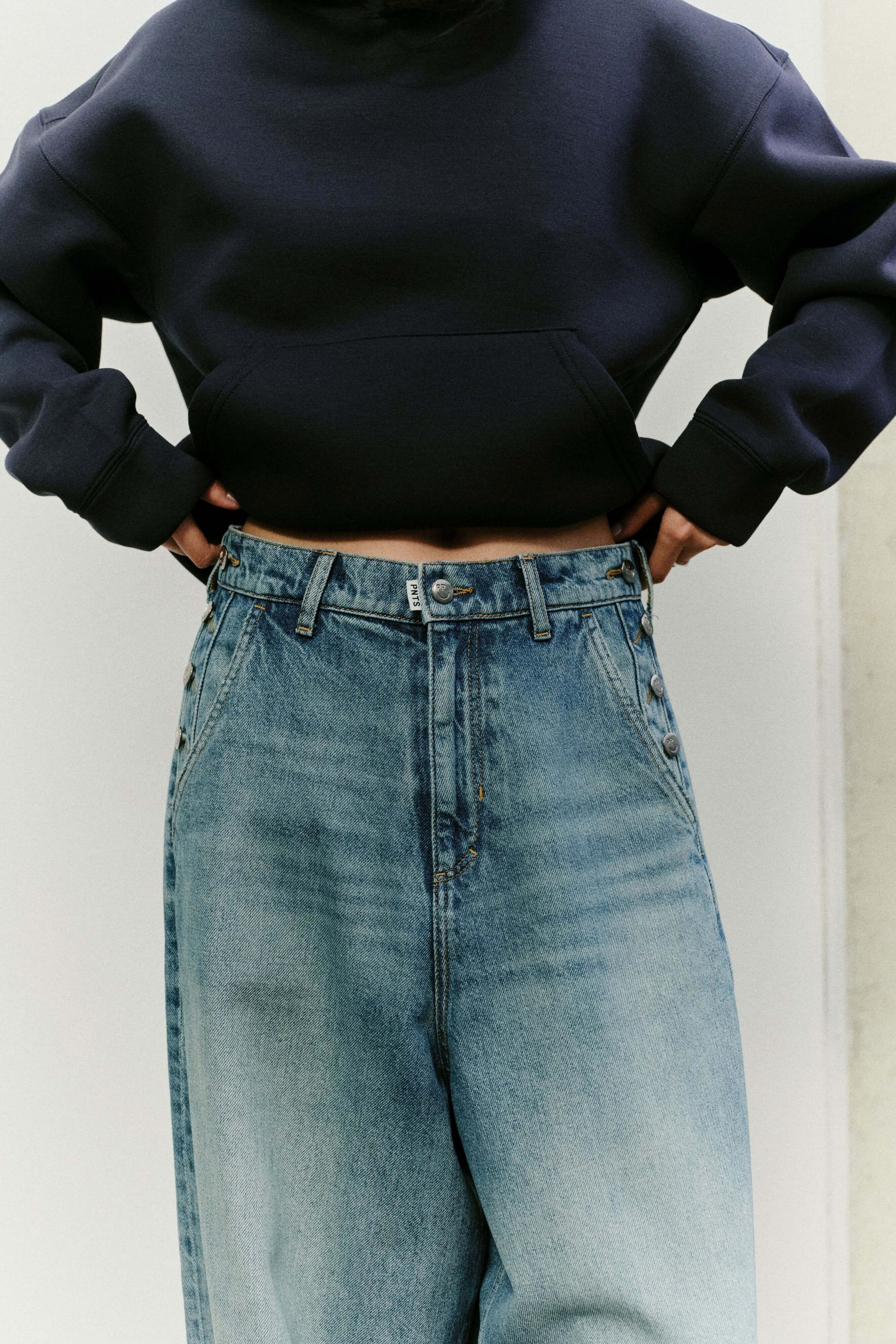 Nahaufnahme des Hosenbunds einer weit geschnittenen Jeans mit dekorativen Knopfdetails an den Taschen und dunkelblauem Hoodie.