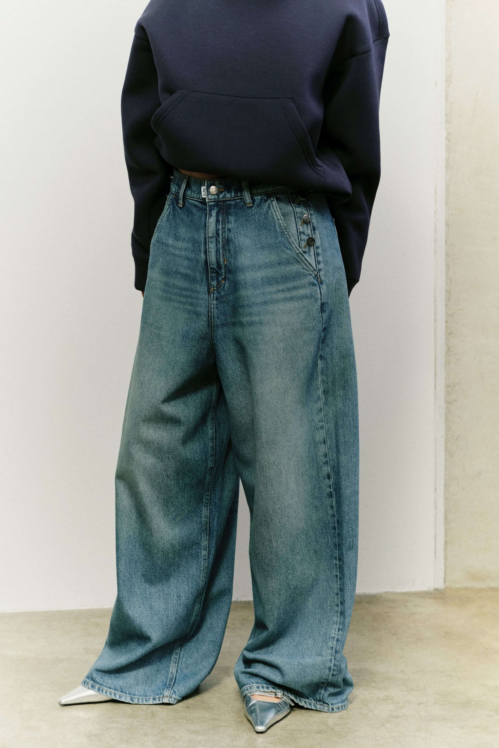 Frau in dunkelblauem Hoodie und extrem weit geschnittener Jeans mit Knopfdetails an den Taschen und silbernen Schuhen.