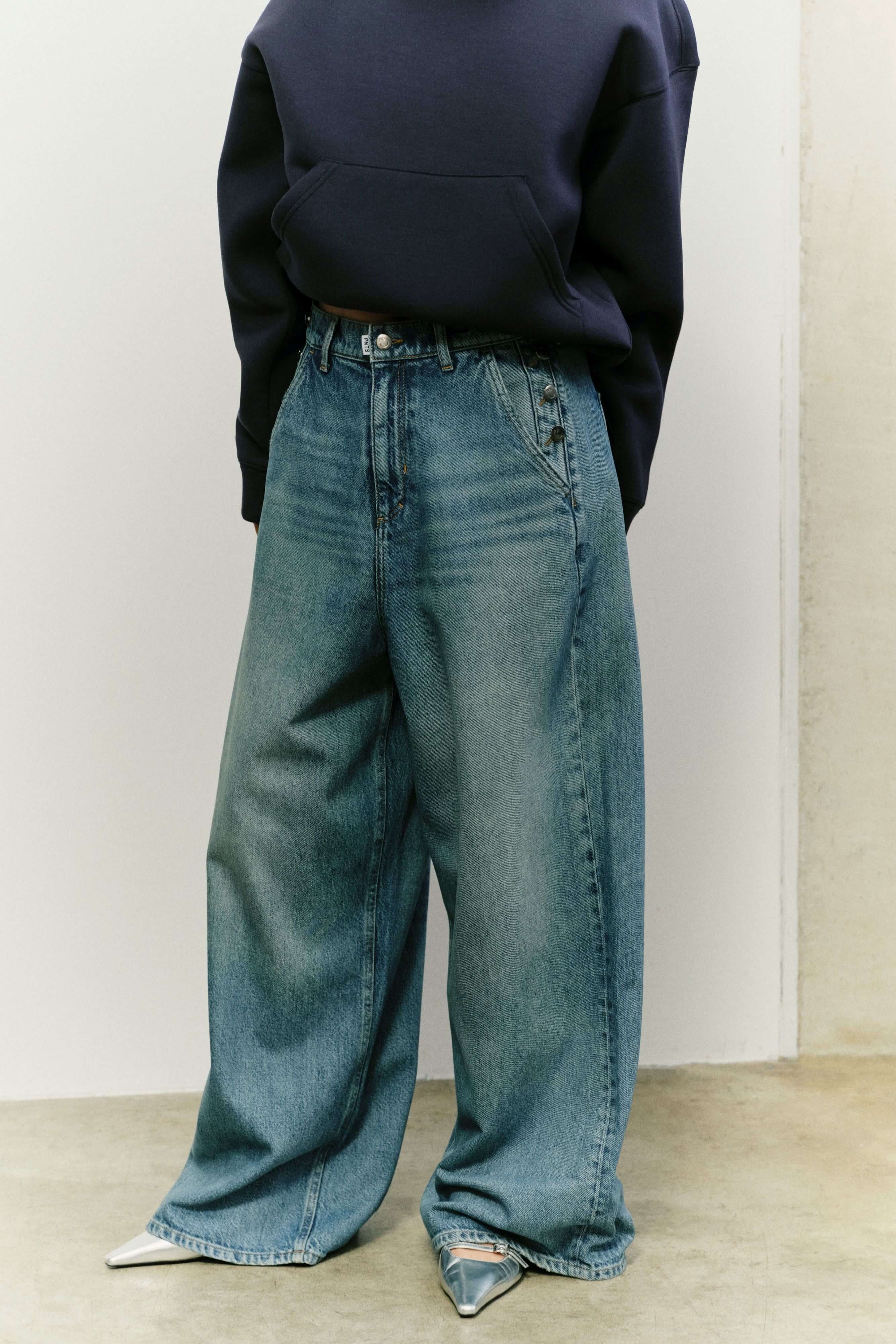 Frau in dunkelblauem Hoodie und extrem weit geschnittener Jeans mit Knopfdetails an den Taschen und silbernen Schuhen.