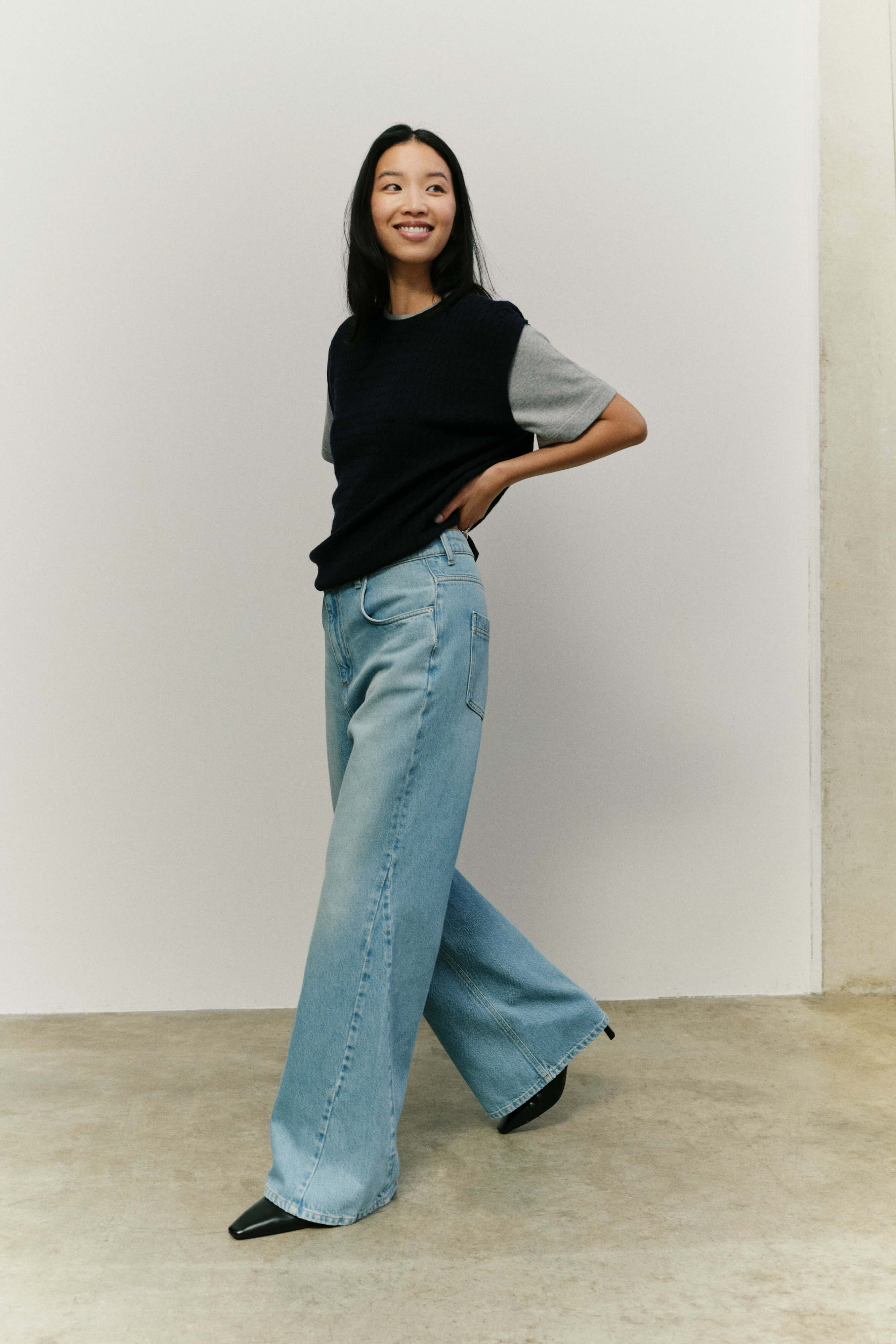 Frau in hellblauer Wide-Leg-Jeans und dunkelblauer Strickweste über einem grauen T-Shirt vor hellem Hintergrund.