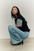 Frau in dunkelblauer Strickweste, grauem T-Shirt und hellblauer Wide-Leg-Jeans in hockender Pose vor hellem Hintergrund.