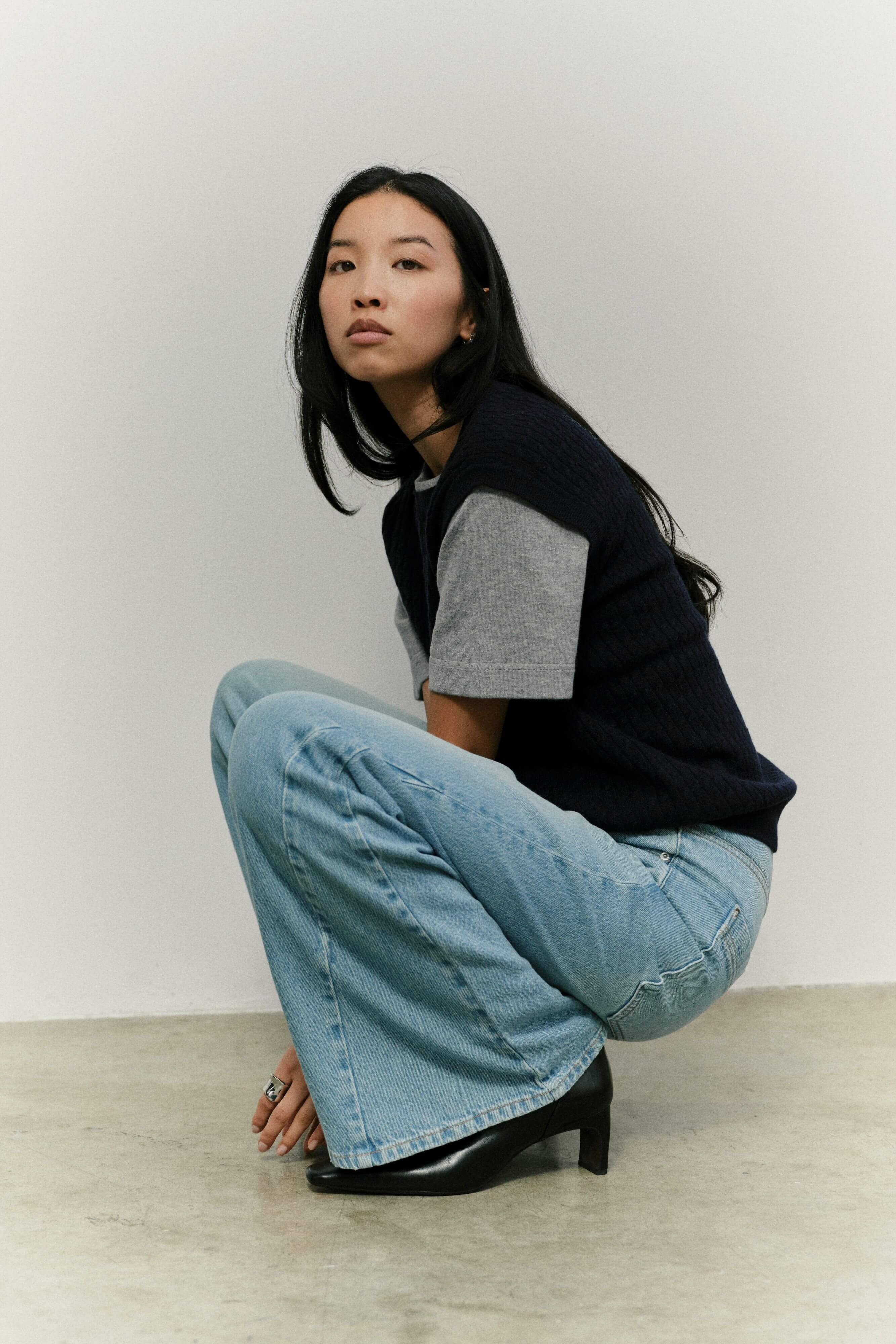 Frau in dunkelblauer Strickweste, grauem T-Shirt und hellblauer Wide-Leg-Jeans in hockender Pose vor hellem Hintergrund.
