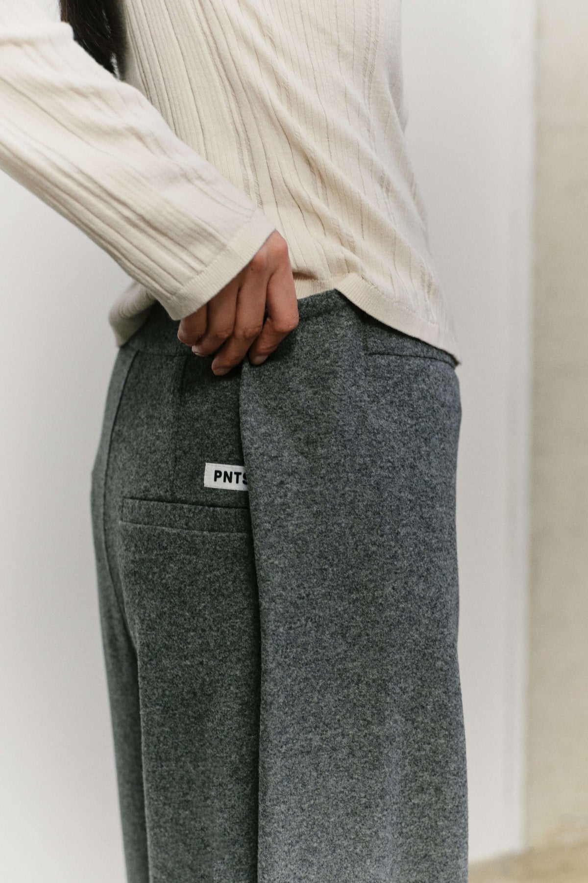 Detailansicht einer grauen melierten Hose mit weißem Logo-Label an der Tasche und hellem Ripp-Oberteil.