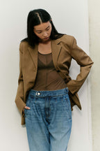 Eine Frau in einem braunen Blazer, einem transparenten braunen Top und einer hellblauen Jeans mit Bundfalten.