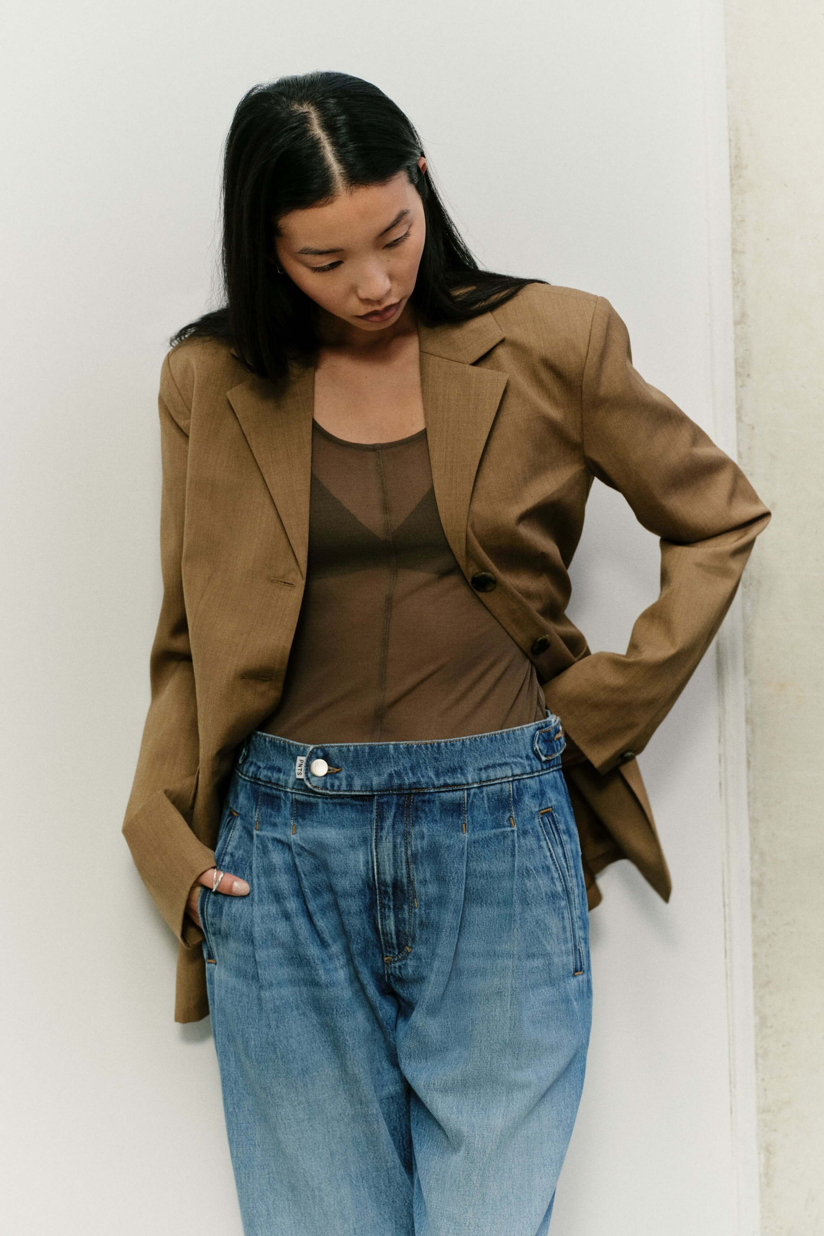 Eine Frau in einem braunen Blazer, einem transparenten braunen Top und einer hellblauen Jeans mit Bundfalten.