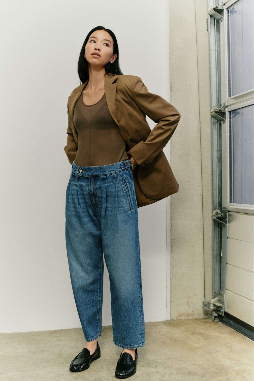 Eine Frau in einem braunen Blazer, transparentem Top, hellblauer Bundfaltenjeans und schwarzen Loafern.