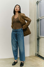 Eine Frau in einem braunen Blazer, transparentem Top, hellblauer Bundfaltenjeans und schwarzen Loafern.