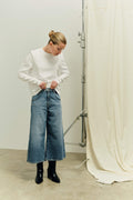 Frau in weißem Langarmshirt und blauer Denim-Culotte mit schwarzen Boots vor hellem Hintergrund.
