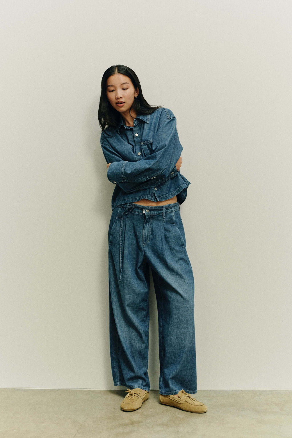 Frau in dunkelblauem All-Denim-Look aus Jeansbluse und weiter Bundfaltenhose mit beigen Mokassins vor hellem Hintergrund.