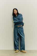Frau in dunkelblauem All-Denim-Look aus Jeansbluse und weiter Bundfaltenhose mit beigen Mokassins vor hellem Hintergrund.