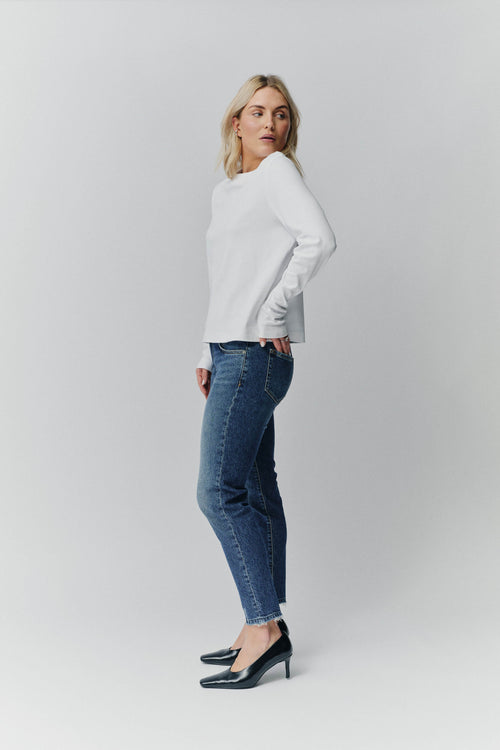 Frau in weißem Langarmshirt und blauer Jeans mit Fransensaum in der Seitenansicht.