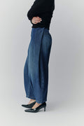 Seitenansicht einer Frau in einem schwarzen Oberteil, einer weiten blauen Jeans-Culotte mit markantem Falten-Detail und schwarzen Pumps.
