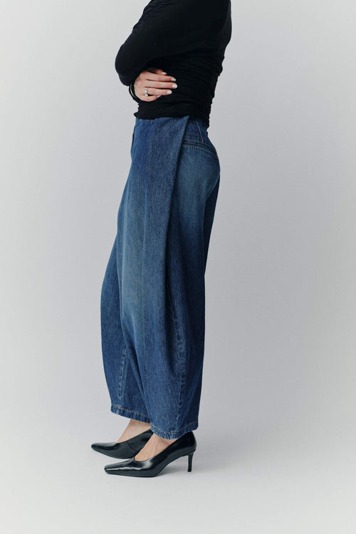Seitenansicht einer Frau in einem schwarzen Oberteil, einer weiten blauen Jeans-Culotte mit markantem Falten-Detail und schwarzen Pumps.