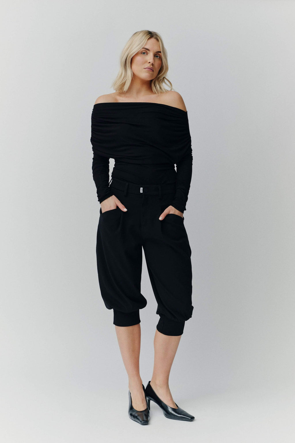 Eine blonde Frau in einem schwarzen schulterfreien Top, einer schwarzen knielangen Ballonhose und schwarzen Pumps vor neutralem Hintergrund.