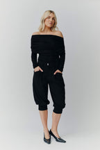 Eine blonde Frau in einem schwarzen schulterfreien Top, einer schwarzen knielangen Ballonhose und schwarzen Pumps vor neutralem Hintergrund.