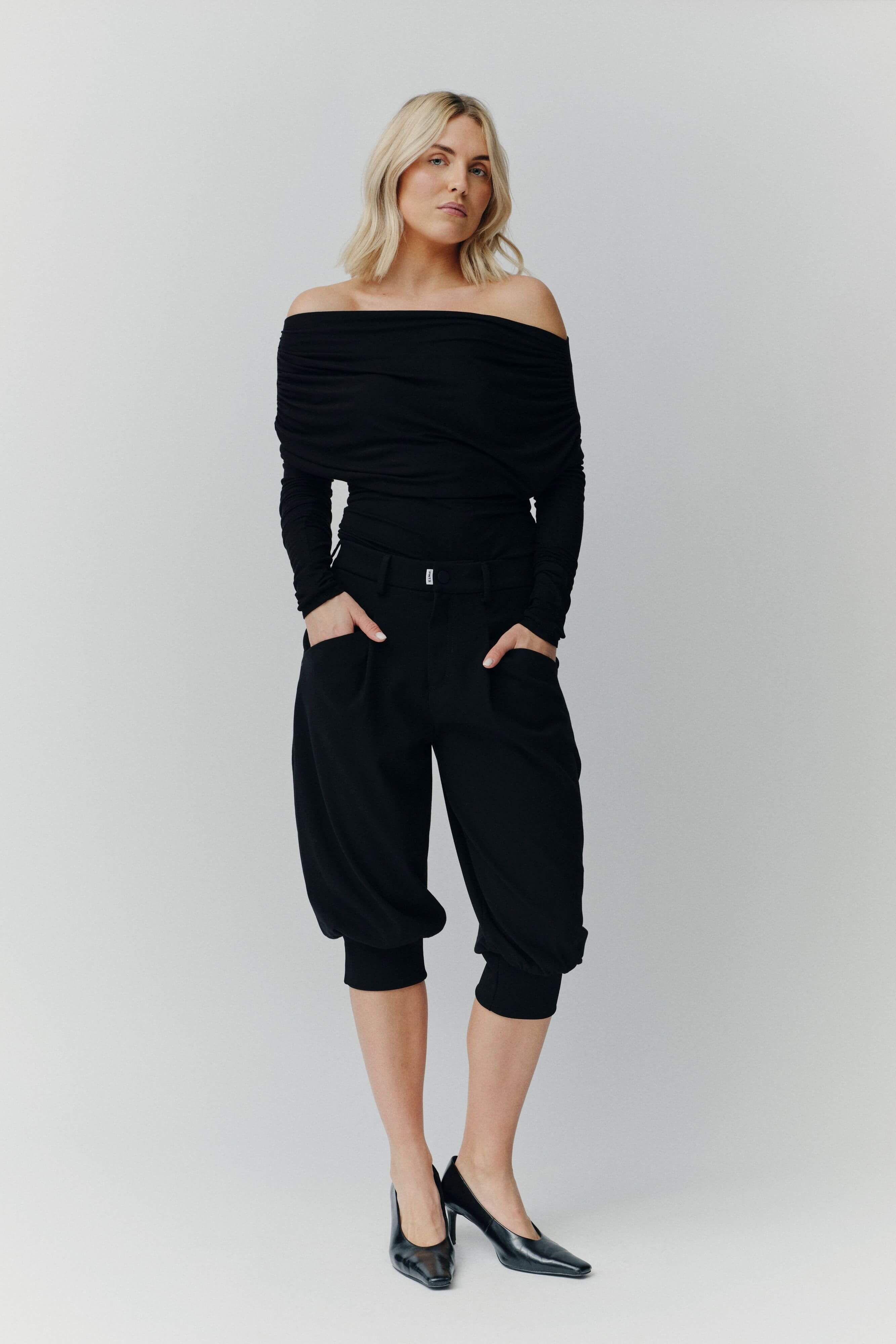 Eine blonde Frau in einem schwarzen schulterfreien Top, einer schwarzen knielangen Ballonhose und schwarzen Pumps vor neutralem Hintergrund.