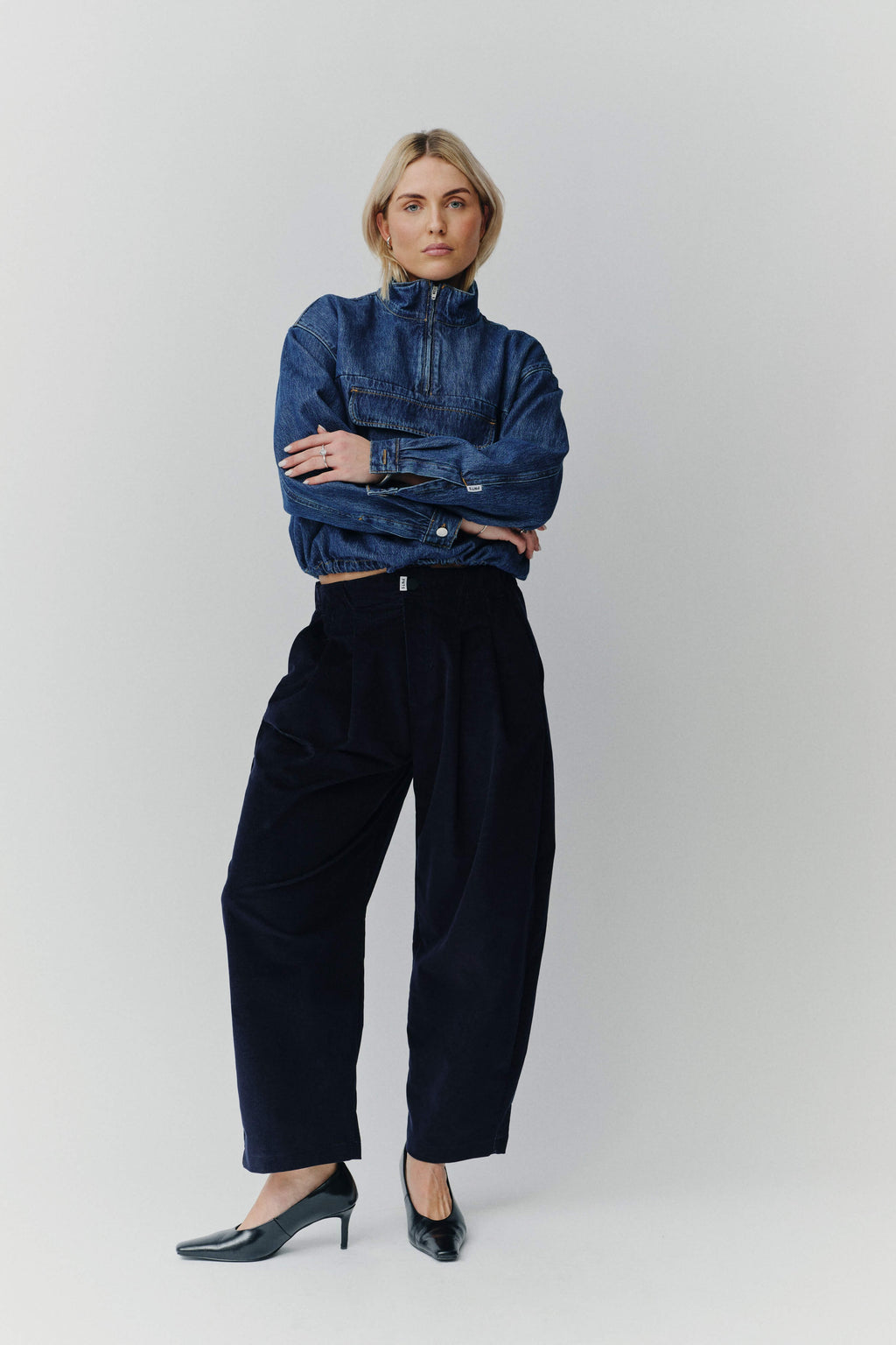 Frau in einem dunkelblauen Denim-Troyer und einer weiten dunkelblauen Cordhose vor hellem Hintergrund.