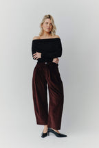 Frau in schwarzem Off-Shoulder-Oberteil und weiter dunkelroter Cordhose vor hellem Hintergrund.