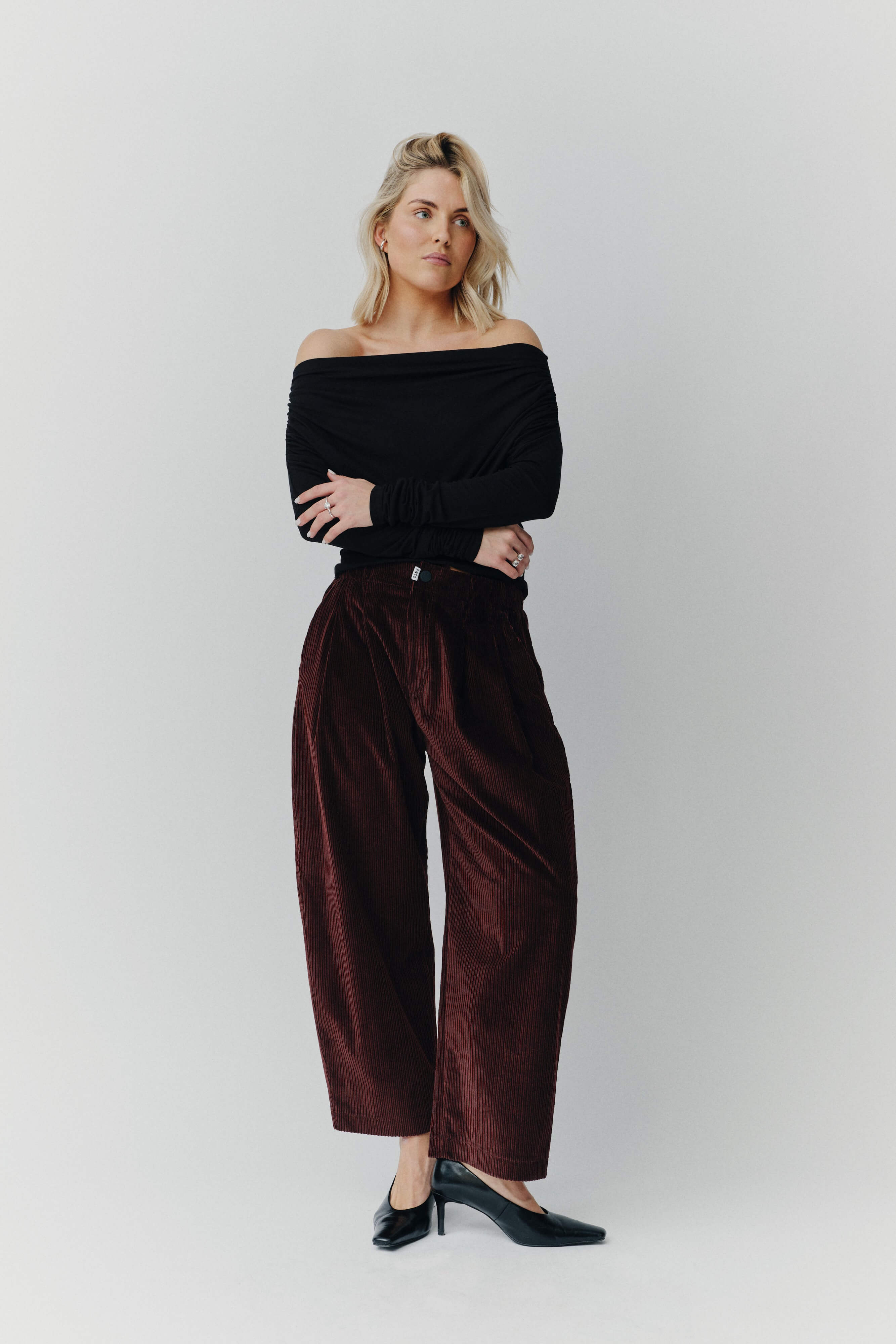 Frau in schwarzem Off-Shoulder-Oberteil und weiter dunkelroter Cordhose vor hellem Hintergrund.
