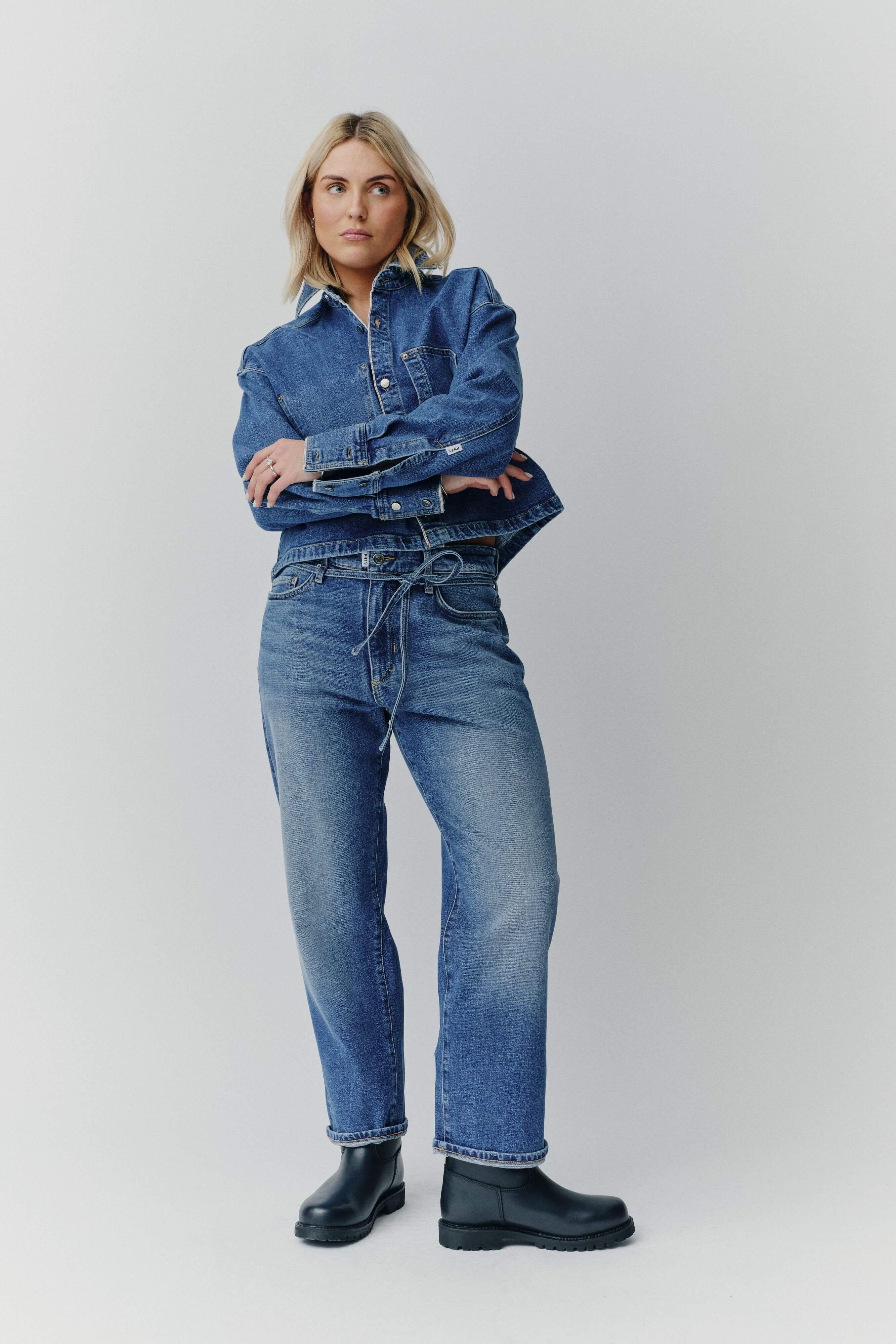 Frau in einem Denim-Outfit mit Jeansjacke, passender Jeans mit Kordelzug und schwarzen Boots vor hellem Hintergrund.