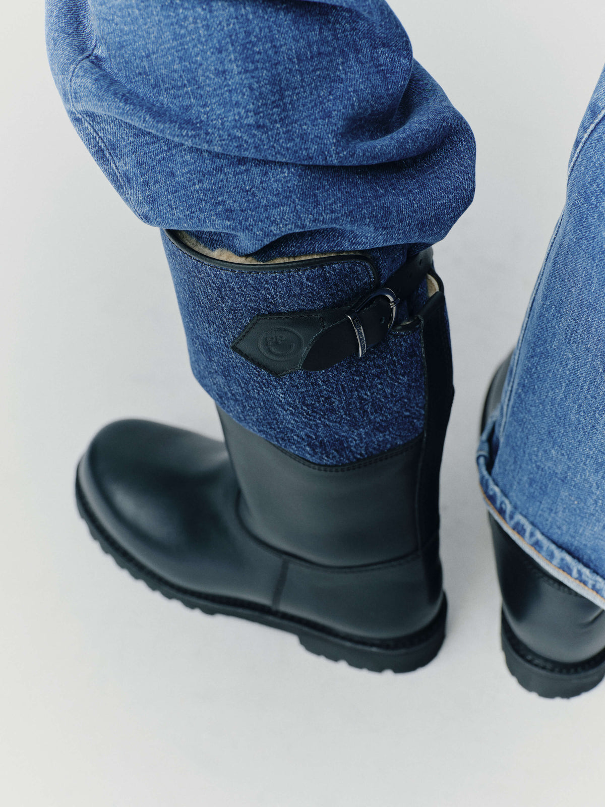 Detailansicht: Schwarzer Lederstiefel mit blauem Schafteinsatz, Schnallenverschluss und sichtbarem Lammfell-Innenfutter, getragen zu Blue Jeans.