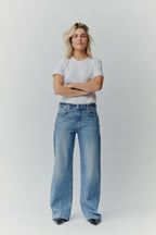 Eine blonde Frau in einem weißen T-Shirt und einer weit geschnittenen blauen Jeans vor neutralem Hintergrund.