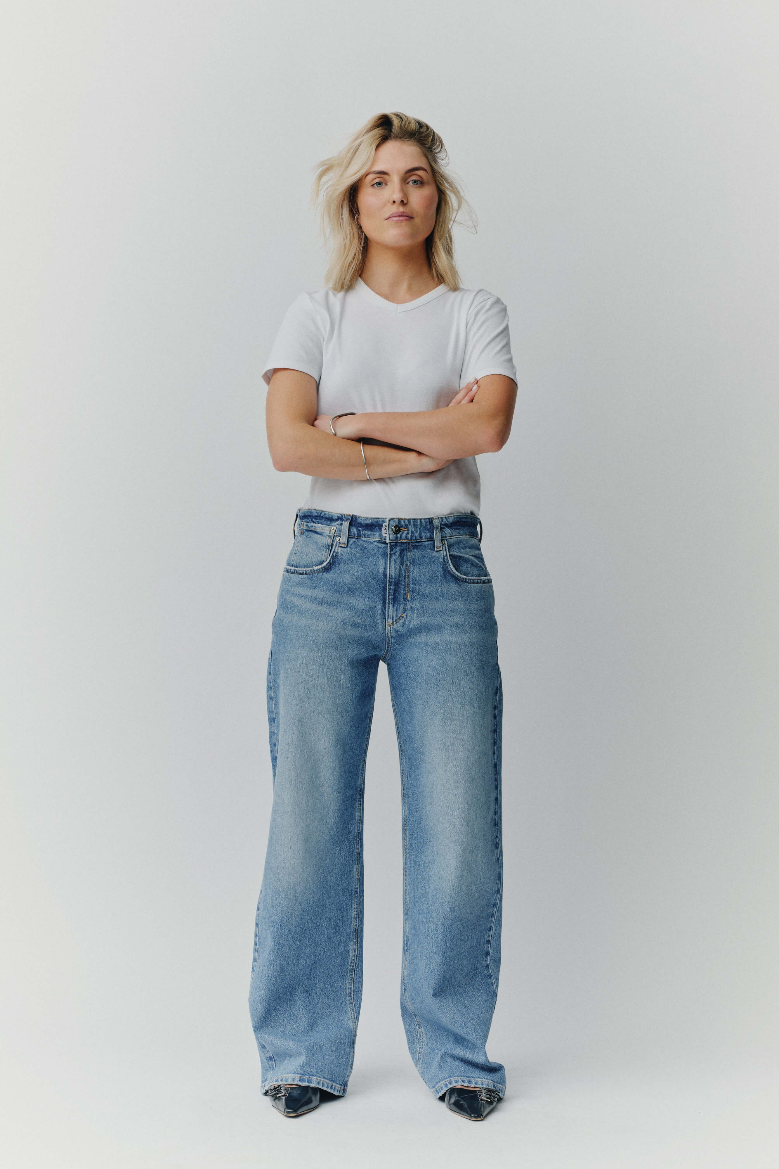 Eine blonde Frau in einem weißen T-Shirt und einer weit geschnittenen blauen Jeans vor neutralem Hintergrund.