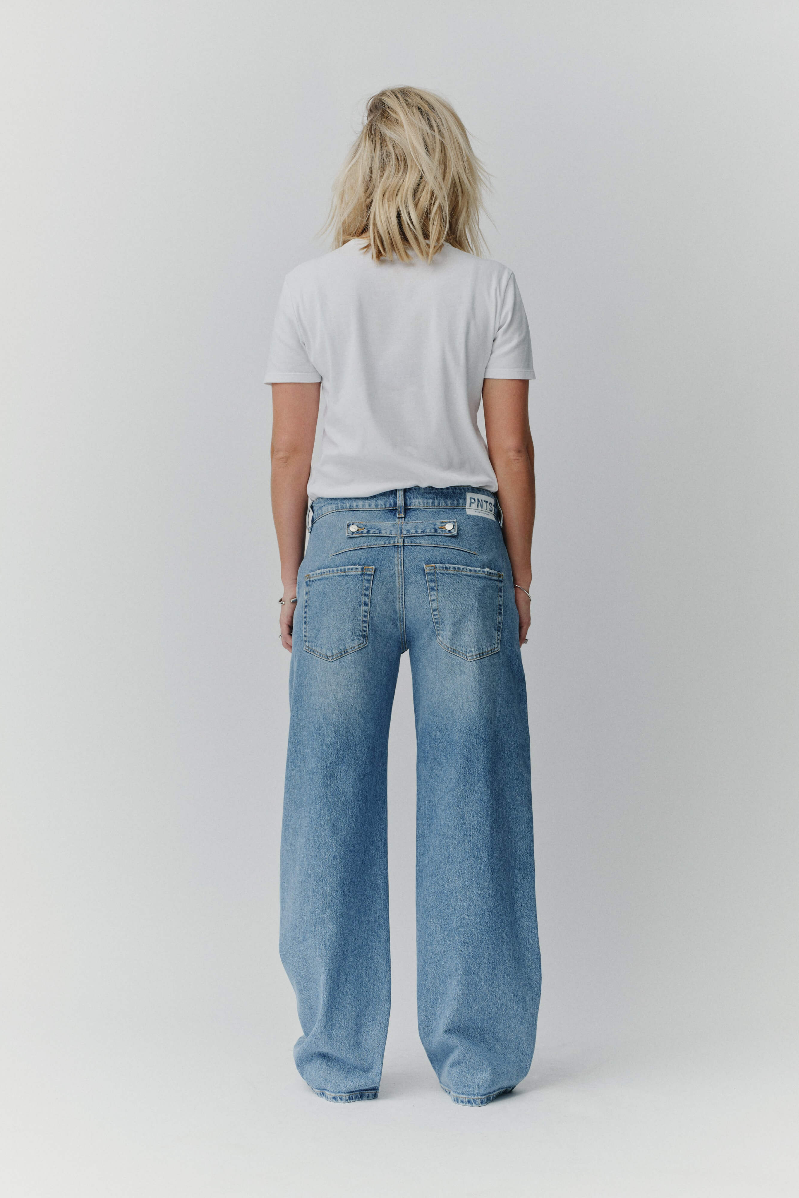 Rückansicht einer Frau in einem weißen T-Shirt und einer weiten blauen Jeans mit PNTS-Label am Bund.