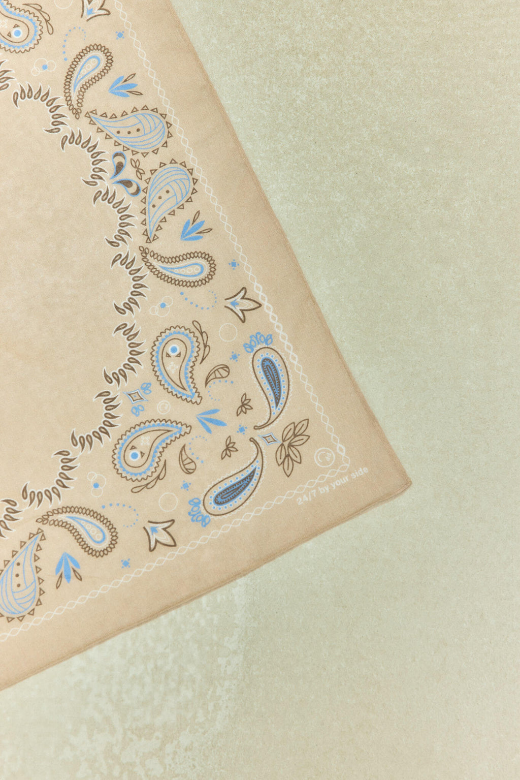 Nahaufnahme eines beigen Bandanas mit blau-braunem Paisley-Muster und dezentem Schriftzug in der Ecke.