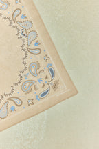 Nahaufnahme eines beigen Bandanas mit blau-braunem Paisley-Muster und dezentem Schriftzug in der Ecke.