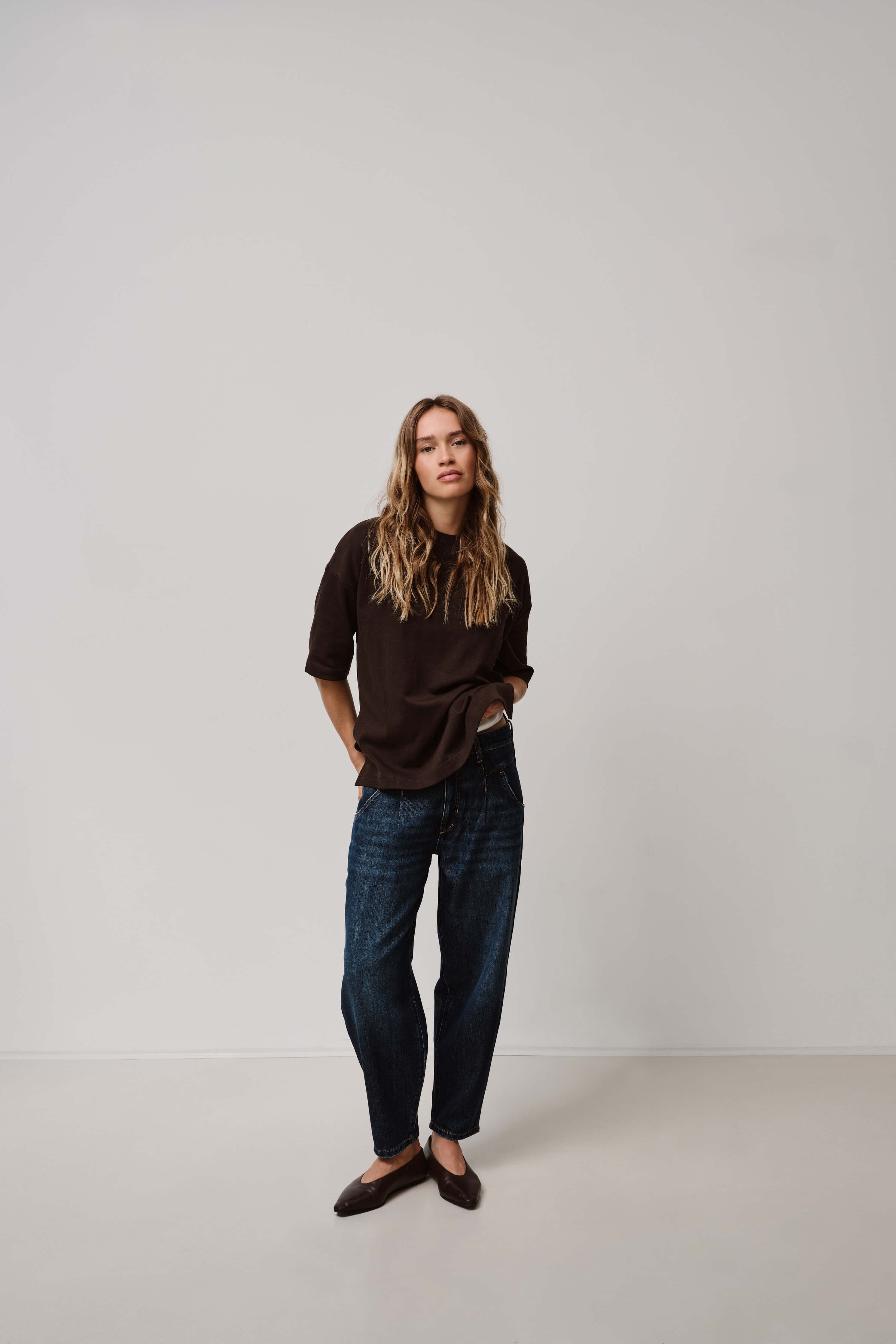 Eine Frau in einem dunkelbraunen Oversize-T-Shirt und einer dunkelblauen Jeans vor neutralem Hintergrund.