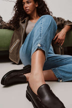 Eine Frau sitzt in einer hellblauen Jeans mit Knopf-Detail am Saum und dunkelbraunen Loafern auf dem Boden.