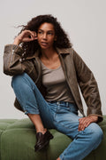 Eine Frau mit lockigen Haaren sitzt in einer braunen Lederjacke, einem grauen Top und hellblauer Jeans auf einem grünen Sofa.