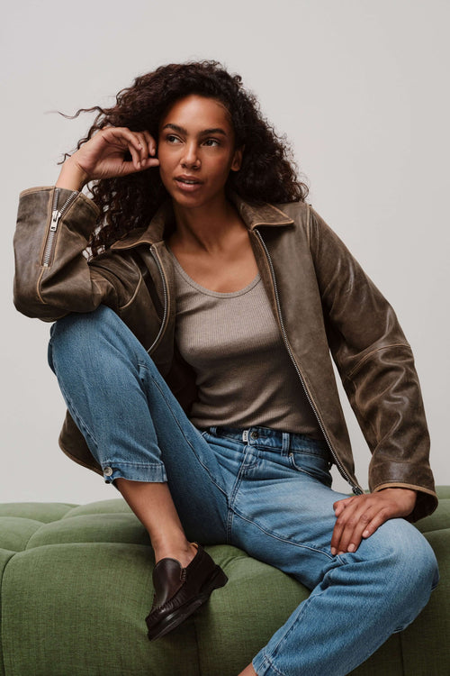 Eine Frau mit lockigen Haaren sitzt in einer braunen Lederjacke, einem grauen Top und hellblauer Jeans auf einem grünen Sofa.