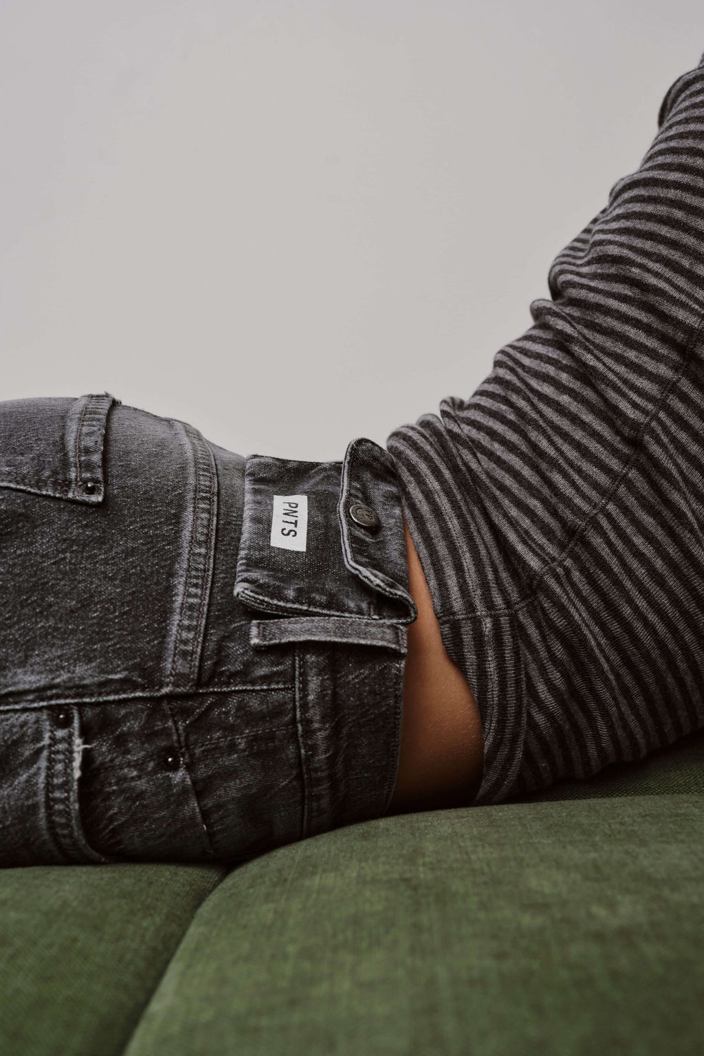 Nahaufnahme des Hosenbunds einer dunkelgrau gewaschenen Jeans mit Logo-Patch und gestreiftem Oberteil.