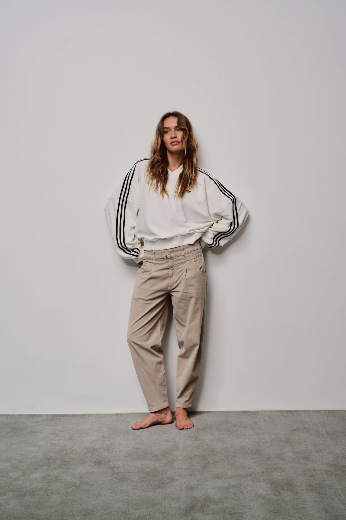Ganzkörperansicht: Model trägt eine locker geschnittene beige Hose mit Bundfalten und ein weißes Sweatshirt.