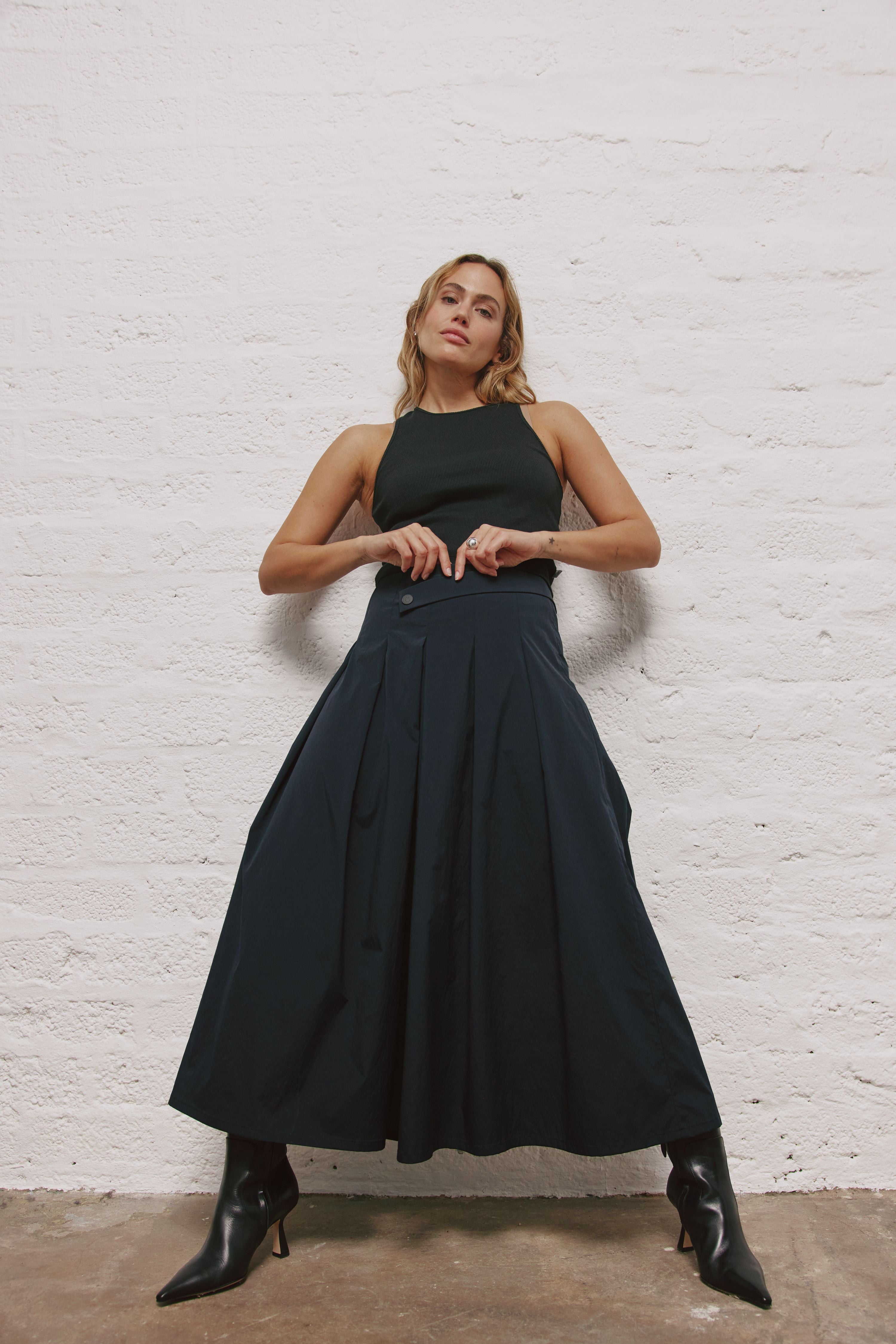 Eine Frau lehnt an einer weißen Wand. Sie trägt schwarze Stiefel, eine blaue, sehr weite Hose und ein schwarzes Top. Ihre Beine sind weit auseinander. Die Hose ist so weit geschnitten, dass es trotz der gespreizten Beine, wie ein Rock aussieht.
