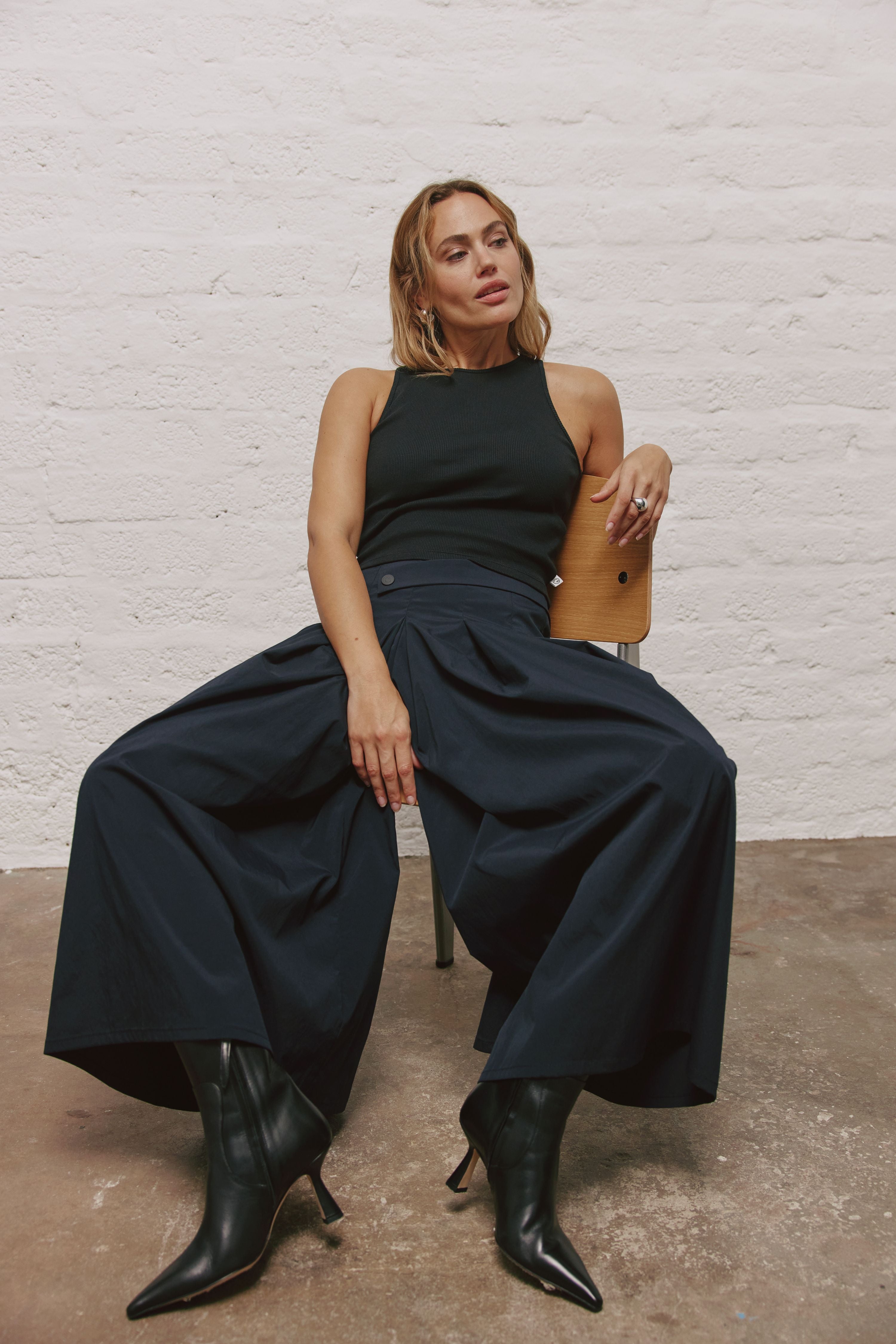Eine Frau sitzt auf einem Stuhl vor einer Wand. Ihr linker Arm ist über der Stuhllehne. Sie trägt schwarze Stiefel, eine sehr weite blaue Hose, die fast wie ein Rock aussieht und ein schwarzes Top.