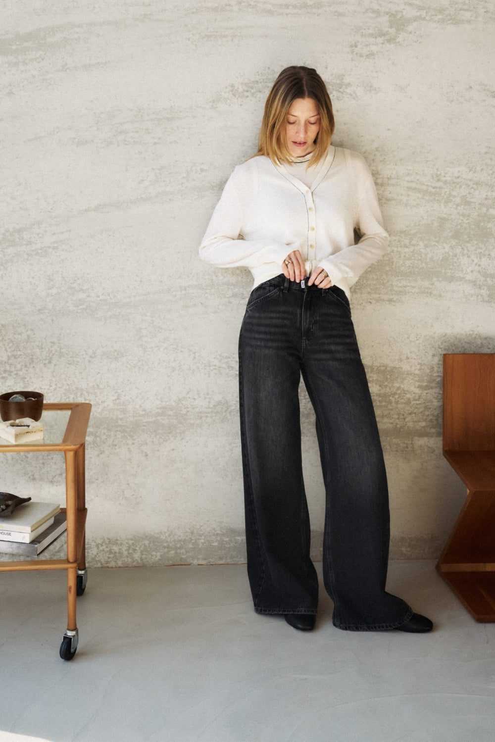 Eine Frau lehnt an einer Wand. Sie schaut auf den Knopf ihrer Hose, den sie gerade schließt. Sie trägt die 16_THE RAVER in dunkelgrau.