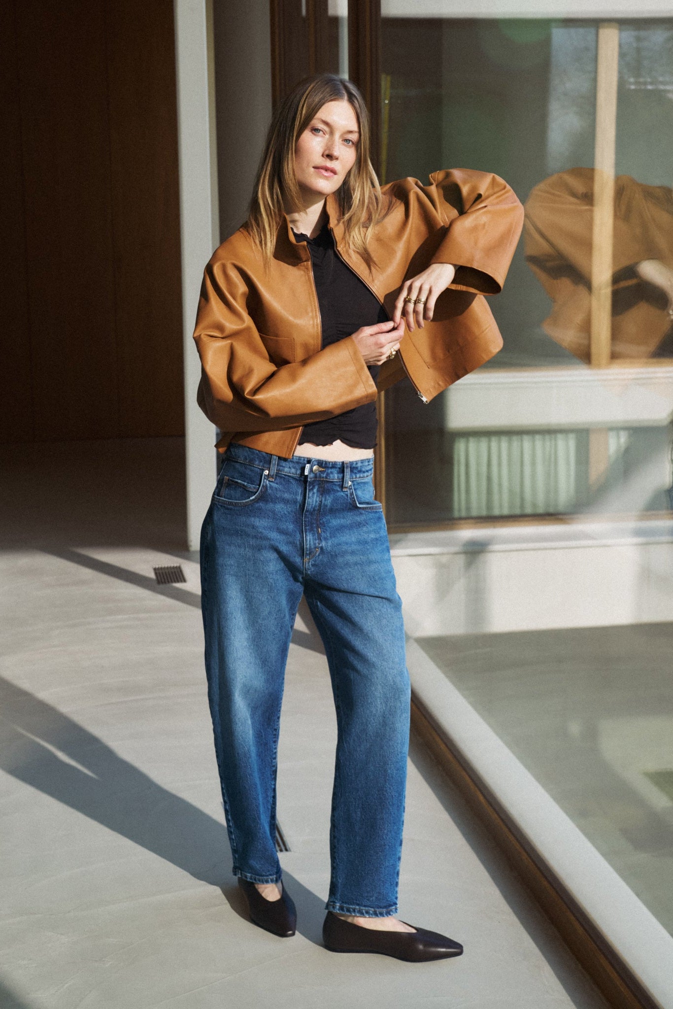 Eine Frau ist an einem Fenster angelehnt und trägt eine rostbraune Lederjacke und dazu eine Jeans 05_THE O SHAPE in dunkelblau. 