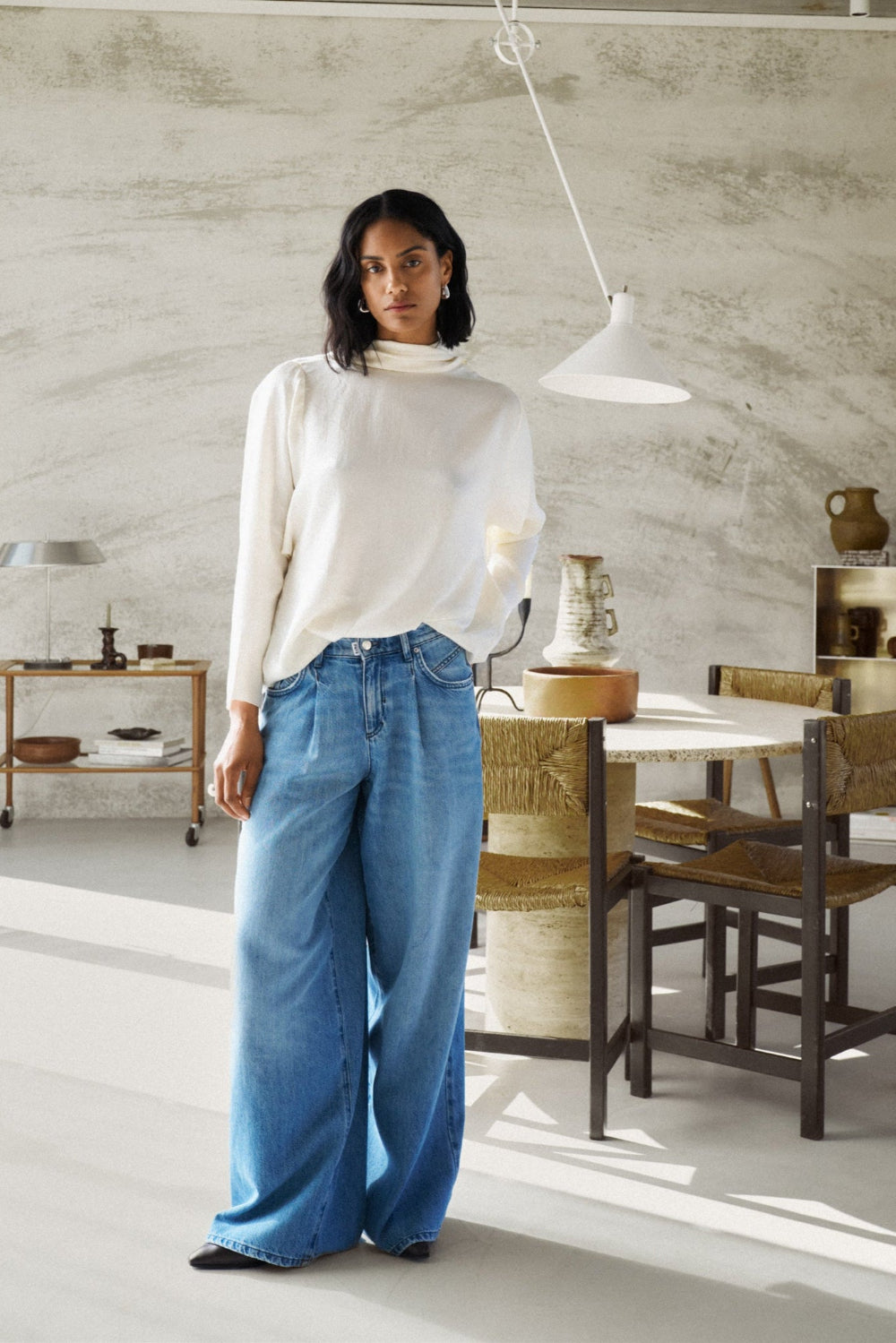 Eine Frau steht vor einem Esstisch und trägt einen weißen Pullover mit einer Jeans 14_THE GENT in hellblau.