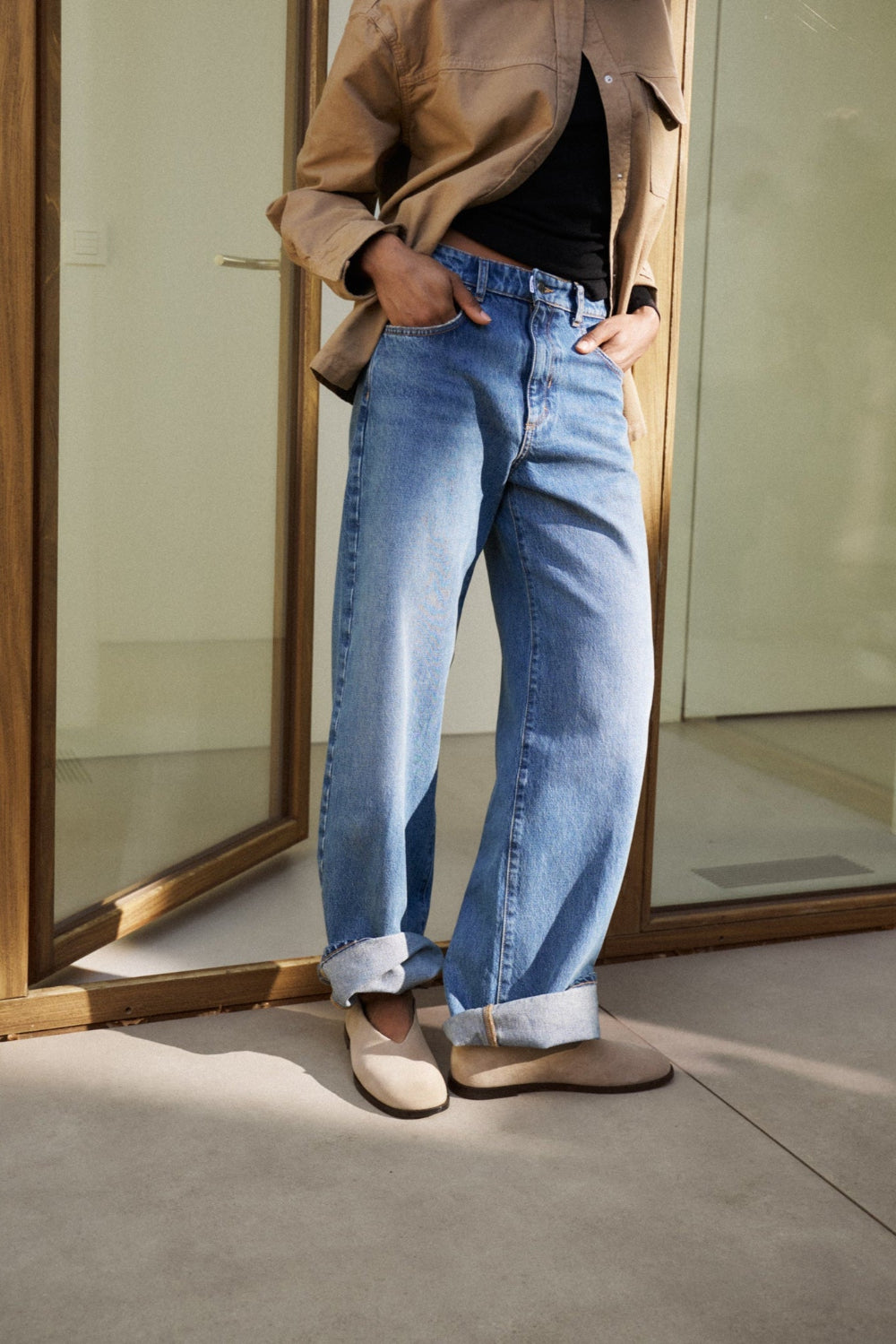 Eine Frau steht angelehnt an einem Fensterrahmen. Sie trägt eine braune Jacke und dazu die Jeans 26_THE LOOSE in mittelblau.