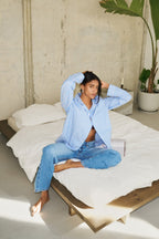 Eine Frau sitzt auf einem Bett und trägt ein blau-weiß gestreiftes Hemd sowie eine Jeans 09_THE BAGGY in hellblau.