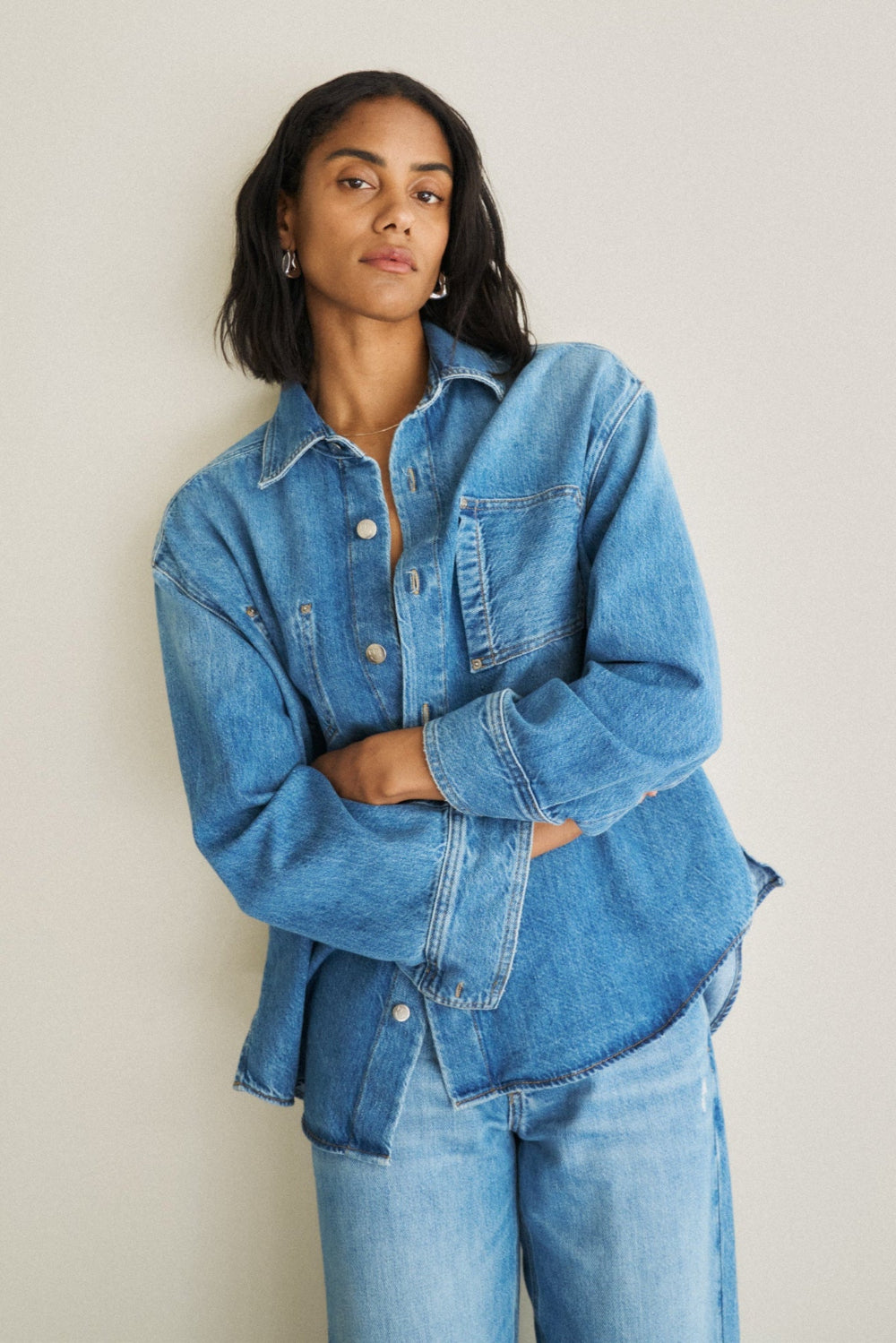 Eine Frau steht vor einer weißen Wand und trägt ein Jeanshemd 27_THE SUIT UP BLOUSE in hellblau.