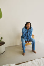 Eine Frau sitzt auf einem Stuhl in einem Raum vor einer weißen Wand und trägt ein Jeanshemd 27_THE SUIT UP BLOUSE in hellblau und eine Jeans 09_THE BAGGY in hellblau dazu.