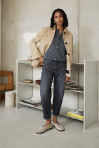 Eine Frau steht vor einem Bücherregal und trägt einen beigen Mantel und dazu eine Jeans 05_THE O SHAPE in dunkelgrau. 