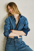 Eine Frau ist angelehnt an einer Wand und trägt ein Jeanshemd mit weißen Streifen. Das Jeanshemd 27_THE SUIT UP BLOUSE STRIPES in mittelblau. 